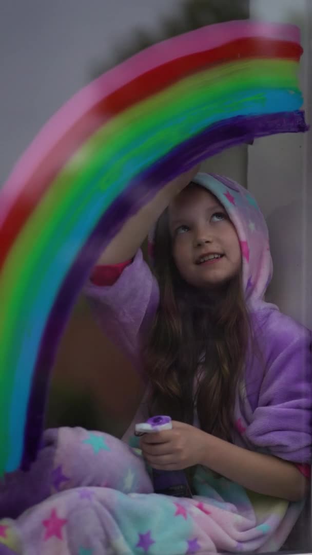 Verticale video. schoolmeisje in pyjama tekent de regenboog op het raam thuis. Blijf thuis, flitsmeute achtervolg de regenboog. — Stockvideo