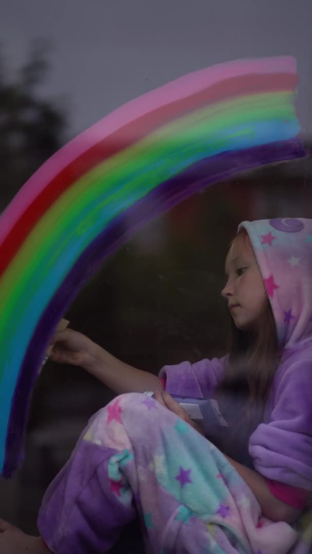 Vídeo vertical. menina da escola em pijama desenha o arco-íris na janela em casa. Fique em casa, flash mob perseguir o arco-íris — Vídeo de Stock