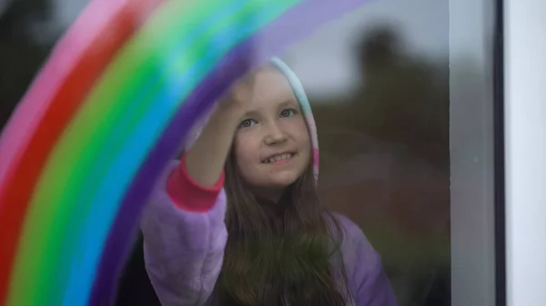 Meisje in pyjama tekent de regenboog op het raam thuis. — Stockfoto