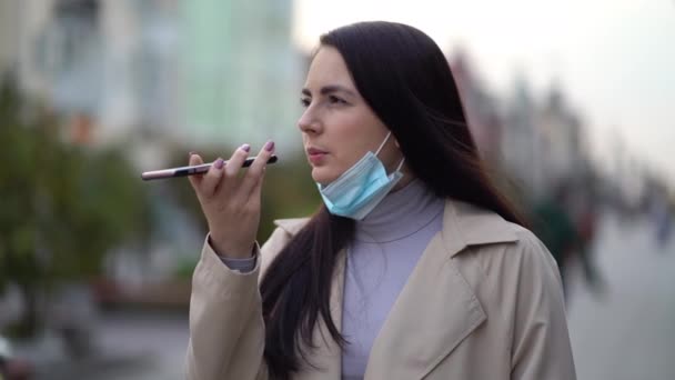 Bonita mujer joven con máscara facial protectora mientras utiliza el teléfono inteligente y el envío de mensajes de voz en la calle urbana — Vídeos de Stock