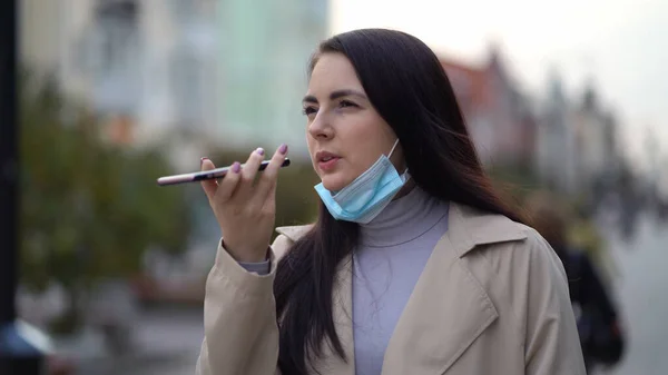 Vacker ung kvinna med skyddande ansiktsmask när du använder smarttelefon och skicka röstmeddelande i den urbana gatan — Stockfoto