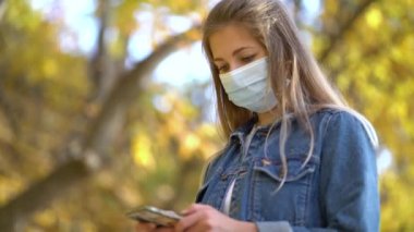 Koruyucu tıbbi maskeli bir kadın sokağa doğru yürüyor. Telefon mesajları kullanıyor. İnternette ikinci dalga covid-19 Coronavirus Virüs Koruma Salgını 'nın haberlerini araştırıyor.