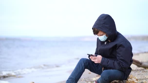 Έφηβος παίζει online βιντεοπαιχνίδια κάθεται στην άδεια παραλία φθινόπωρο κατά τη διάρκεια της πανδημίας εποχή coronavirus — Αρχείο Βίντεο