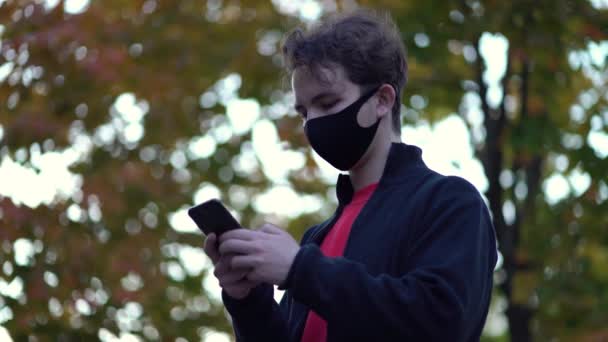 Portret chłopca w ochronnej masce medycznej idzie na ulicę używa tekstów telefonicznych przewija się po internecie szukając wiadomości o drugiej fali covid-19 pandemii ochrony przed wirusami koronawirusowymi — Wideo stockowe