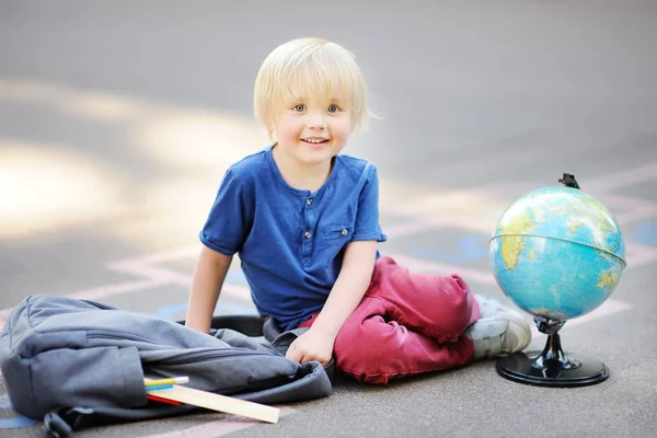 Cute blond boy doing homework sitting on school yard after school with bags laying near. Back to school concept. Little boy on school yard