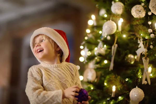 小男孩戴着圣诞老人的帽子准备庆祝圣诞节 逗人喜爱的孩子装饰圣诞树与紫色玻璃玩具 — 图库照片