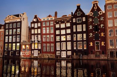 Alacakaranlıkta Amsterdam'da Damrak kanalının ünlü dans evleri ve Amstel nehri suyunda yansımaları