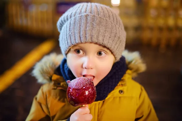 赤いリンゴを食べる男の子はクリスマス マーケットにキャラメルで覆われています 伝統的な子供の楽しさと楽しいクリスマスの時間の間に 子供とお菓子 — ストック写真