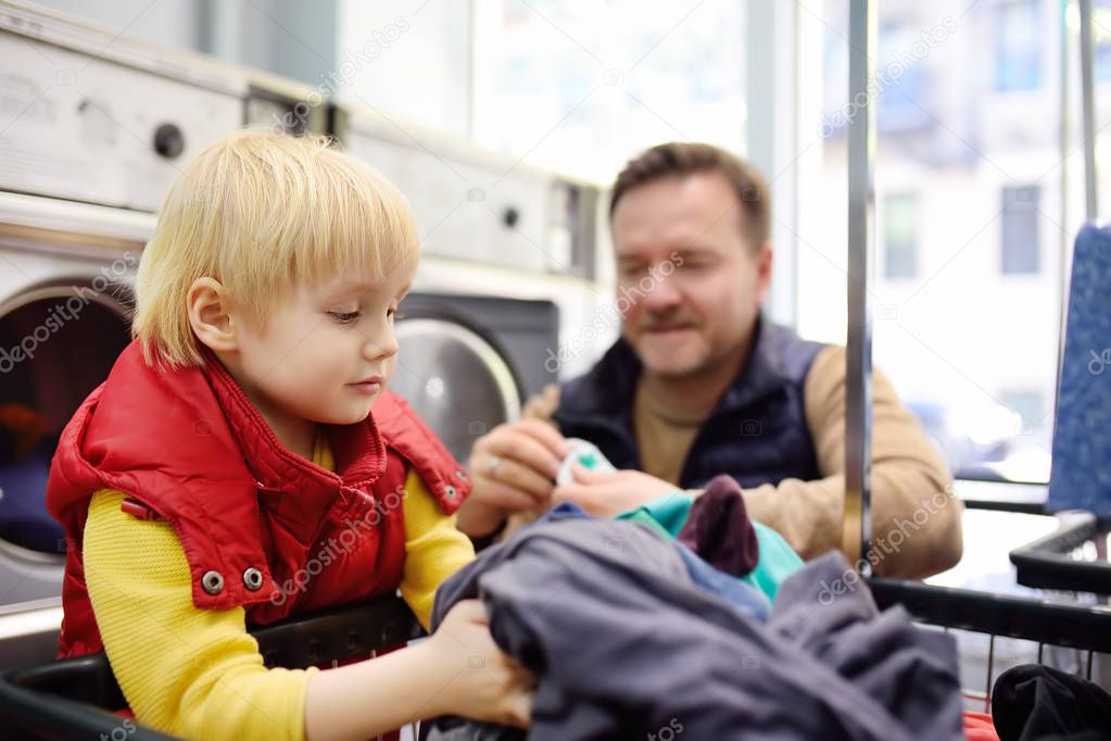 A little boy loads clothes into the washing machine in public laundrette. Family in public laundry