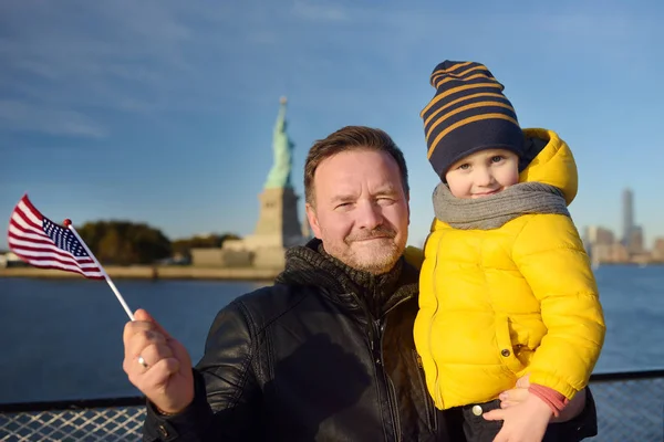 父と彼の幼い息子の自由の女神像の背景にアメリカの国旗を保持しています 家族旅行のコンセプト — ストック写真