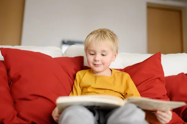Little boy is sitting at home on the couch and reading a book. Learning to read. Home education concept.
