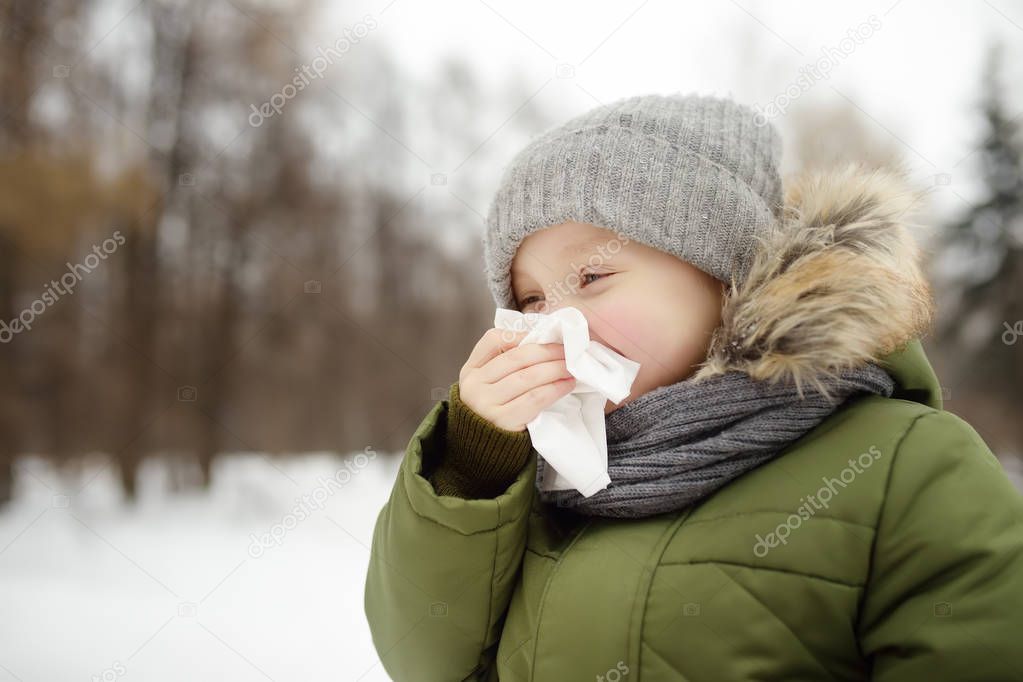 Little boy sneezing and wipes nose with napkin during walking in winter park. Flu season and cold rhinitis. Allergic kid. Sick kids.