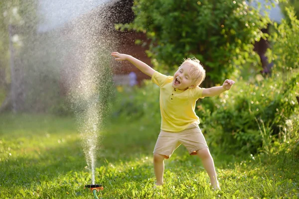 日当たりの良い家の裏庭に 庭のスプリンクラーで遊んで面白い少年 未就学児の子は 水のスプレーを楽しんでします 子供のための夏の屋外活動 — ストック写真