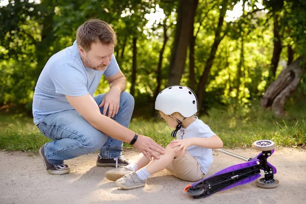 Little boy in safety helmet falls during learning to ride scooter. Father comforting his son after crash. Active family leisure. Child in helmet. Safety, sports, leisure with kids