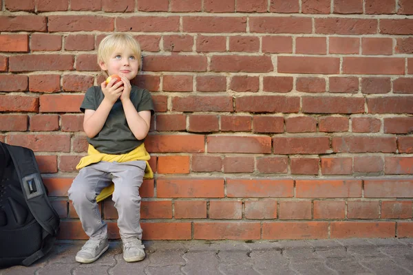 Student with backpack sat down to eat apple near the school building. School meals. — Stock Photo, Image