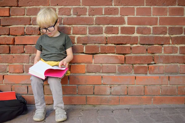 학교 건물 근처에서 휴식을 취하며 숙제를 하고 있는 안경글한 꼬마 소년. 학교 개념으로 돌아가기. — 스톡 사진