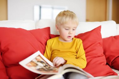 Küçük çocuk evde kanepede oturuyor ve kitap okuyor. Okumayı öğrenme.