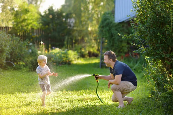 Anak kecil yang lucu dengan ayahnya bermain dengan selang taman di halaman belakang yang cerah. Anak prasekolah bersenang-senang dengan semprotan air . — Stok Foto