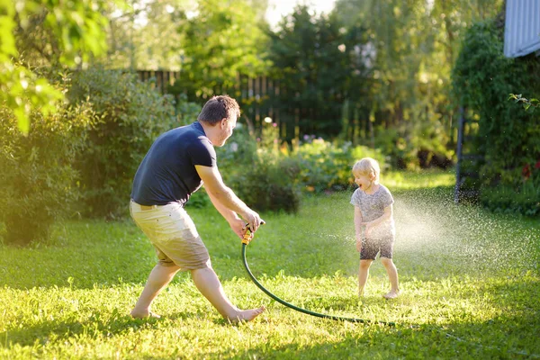 Anak kecil yang lucu dengan ayahnya bermain dengan selang taman di halaman belakang yang cerah. Anak prasekolah bersenang-senang dengan semprotan air . — Stok Foto