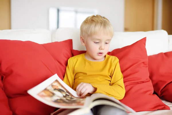 Little boy is sitting at home on the couch and reading a book. Learning to read. — Stock Photo, Image