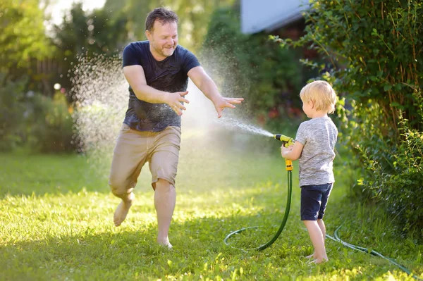 Engraçado menino com seu pai brincando com mangueira de jardim no quintal ensolarado. Criança pré-escolar se divertindo com spray de água . — Fotografia de Stock