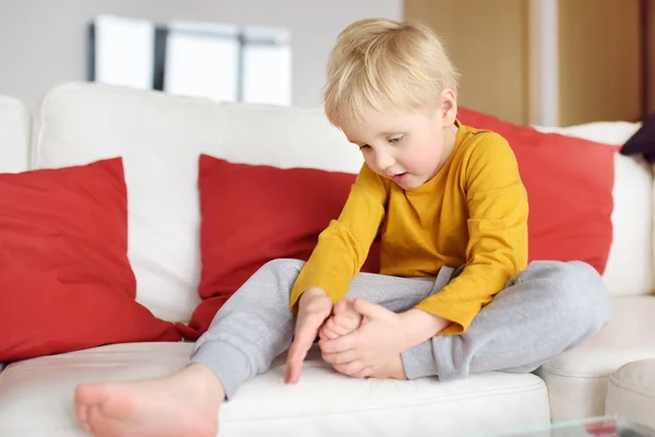 Le petit garçon examine la jambe endolorie assise sur le canapé à l'intérieur. Traumatisme, ecchymoses, éclats . — Photo