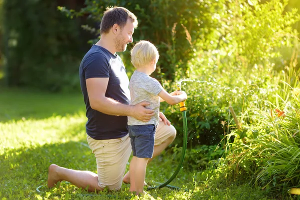 Engraçado menino com seu pai brincando com mangueira de jardim no quintal ensolarado. Criança pré-escolar se divertindo com spray de água . — Fotografia de Stock