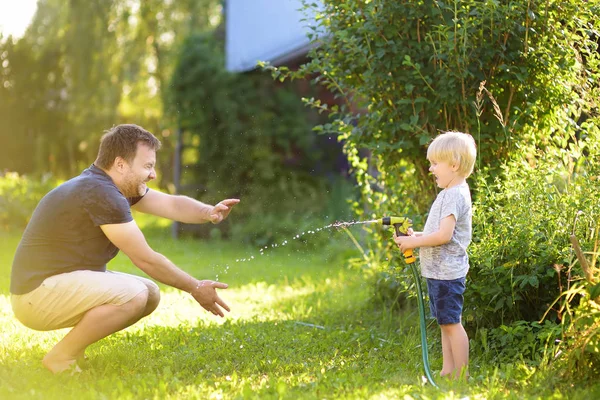 Engraçado menino com seu pai brincando com mangueira de jardim no quintal ensolarado. Criança pré-escolar se divertindo com spray de água . — Fotografia de Stock