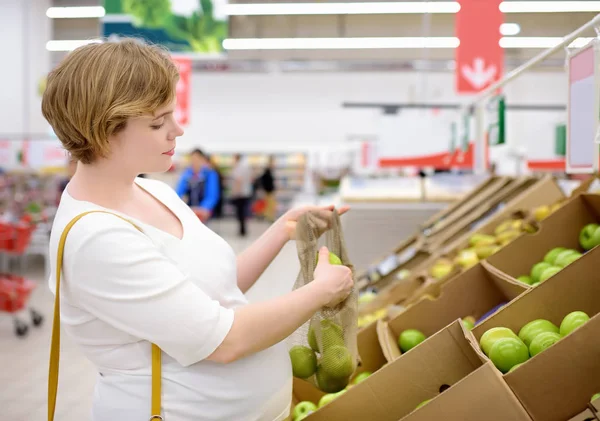 Mujer embarazada joven eligiendo frutas y verduras en bolsa de compras de malla en el supermercado —  Fotos de Stock