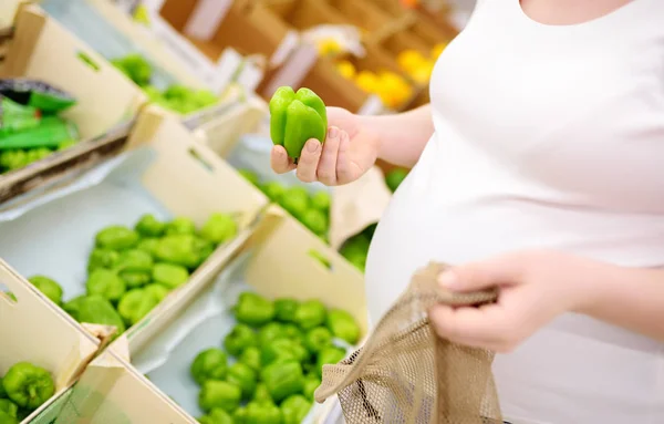 Jovem grávida escolhendo frutas e legumes em malha saco de compras no supermercado — Fotografia de Stock