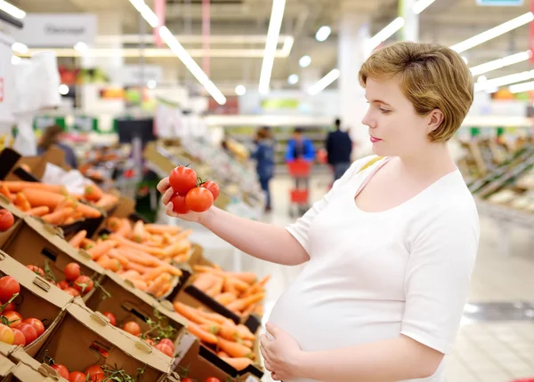 Young pregnant woman choosing fresh tomato in supermarket — Stock Photo, Image