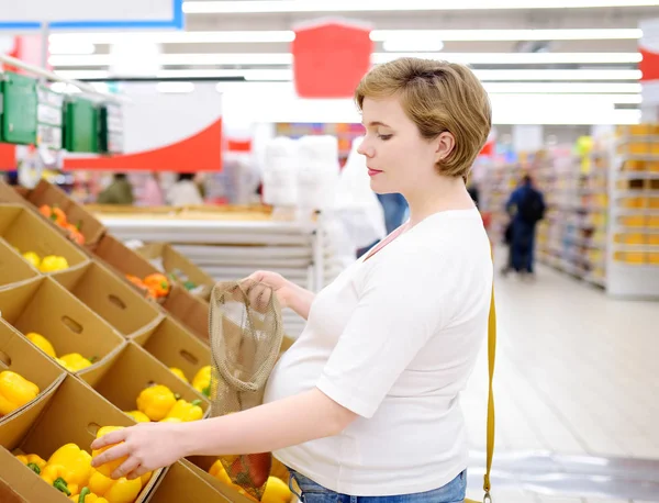 Mujer embarazada joven eligiendo frutas y verduras en bolsa de compras de malla en el supermercado — Foto de Stock