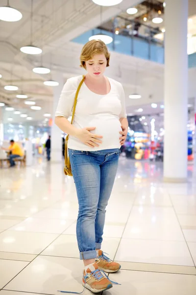 Young pregnant woman in despair because of the untied laceup in the hall of the shopping center. Problems of pregnant women with big belly lace up shoes.