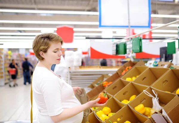Young pregnant woman choosing fruits and vegetables in mesh shopping bag in supermarket — Stock Photo, Image