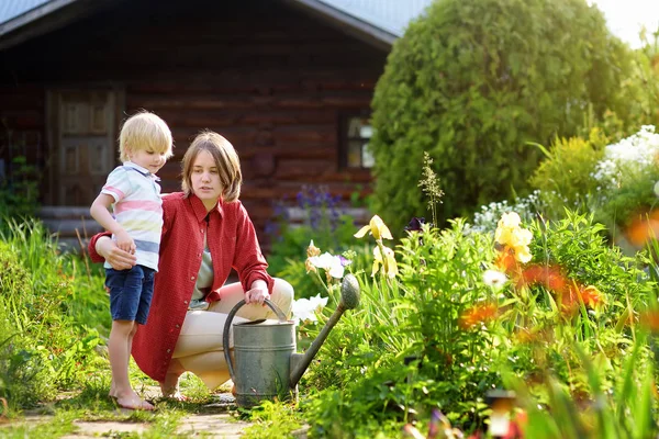 かわいい小さな男の子と彼の若い母親は、夏の晴れた日に庭で植物に水をやります — ストック写真