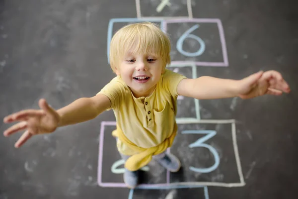 Pequeño niño en el azadón dibujado en asfalto abrazarte. Vista desde arriba. Niño jugando hopscotch juego en el patio al aire libre en un día soleado . —  Fotos de Stock