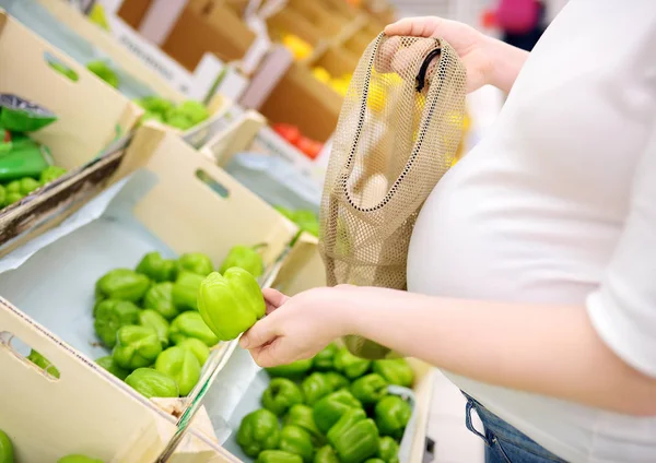 Junge schwangere Frau wählt Obst und Gemüse in Maschen-Einkaufstasche im Supermarkt — Stockfoto