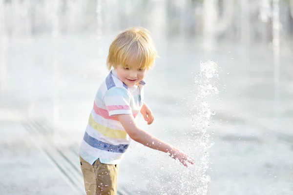 小さな男の子は晴れた夏の日に噴水のウォータージェットの間の広場で遊ぶ. — ストック写真