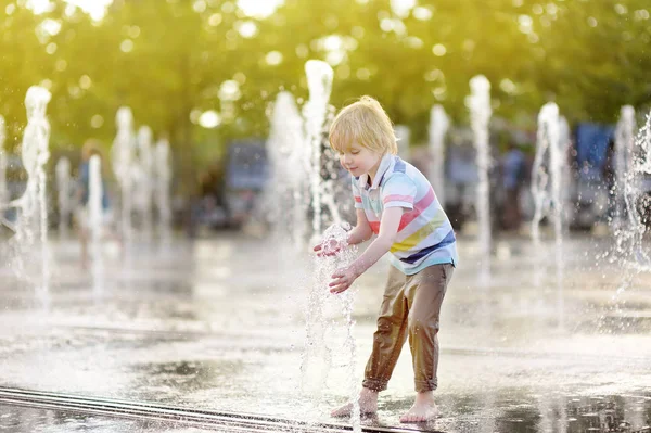 小さな男の子は晴れた夏の日に噴水のウォータージェットの間の広場で遊ぶ. — ストック写真