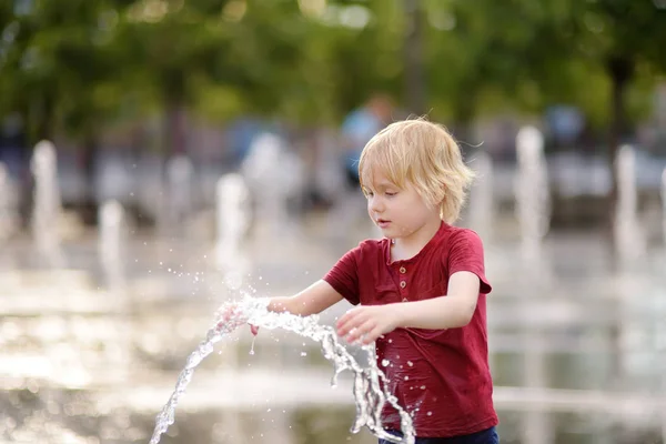 Niño pequeño juega en la plaza entre los chorros de agua en la fuente en el soleado día de verano . —  Fotos de Stock