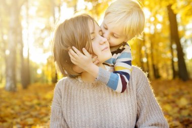Küçük çocuk annesini kucaklıyor ve güneşli sonbahar parkında yürüyüş sırasında onu öpüyor.