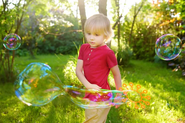 Милый маленький мальчик играет с большими пузырьками на открытом воздухе. Ребенок раздувает большие и маленькие пузырьки одновременно . — стоковое фото
