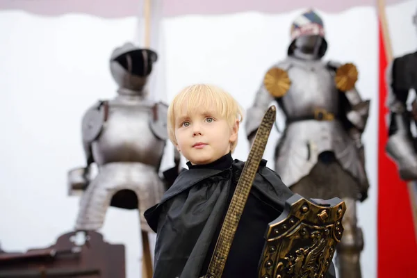 Retrato de um menino bonito vestido como um cavaleiro medieval com uma espada e um escudo no fundo da armadura do cavaleiro . — Fotografia de Stock