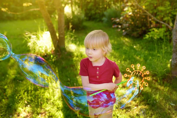 Cute little boy is playing with big bubbles outdoor. Child is blowing big and small bubbles simultaneously. — Stock Photo, Image