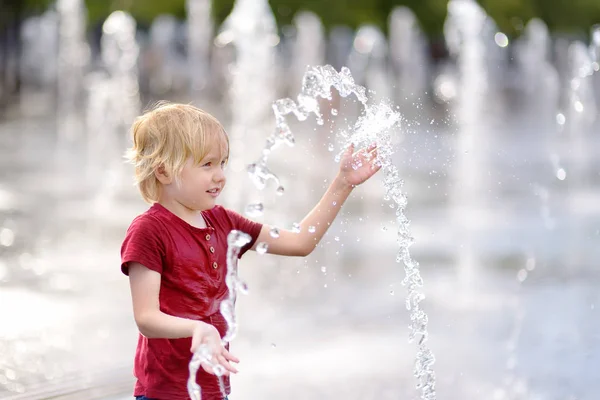 小さな男の子は晴れた夏の日に噴水のウォータージェットの間の広場で遊ぶ. — ストック写真