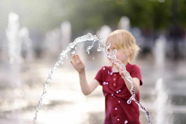 小さな男の子は晴れた夏の日に噴水のウォータージェットの間の広場で遊ぶ。水のジェットに焦点を当てる. — ストック写真