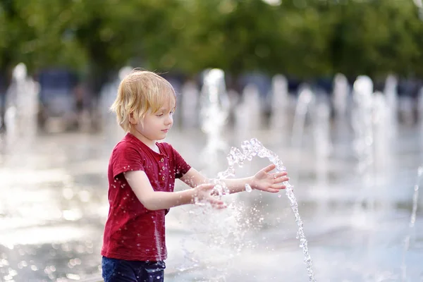 小さな男の子は晴れた夏の日に噴水のウォータージェットの間の広場で遊ぶ. — ストック写真