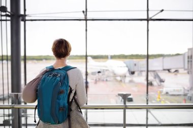 Bir sırt çantası ve seyahat yastığı ile kadın havaalanının bekleme alanında pencerede uçakta görünüyor.