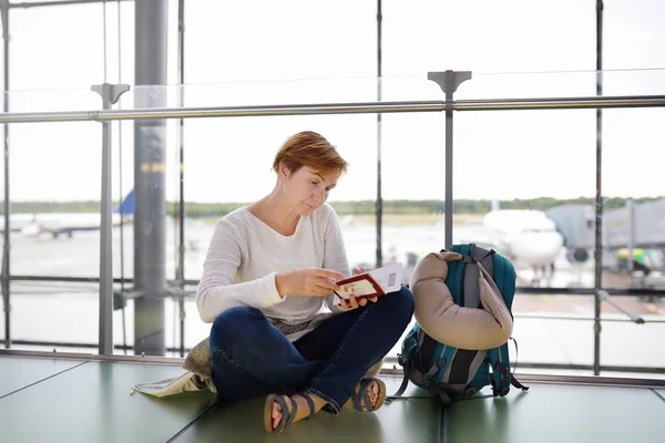 Зріла жінка з рюкзаком і проїзною подушкою, що сидить в зоні очікування аеропорту і перевіряє її квиток . — стокове фото