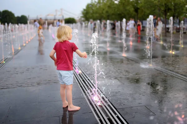 Little boy plays in the square between the water jets in the fountain at summer evening. — Stock Photo, Image